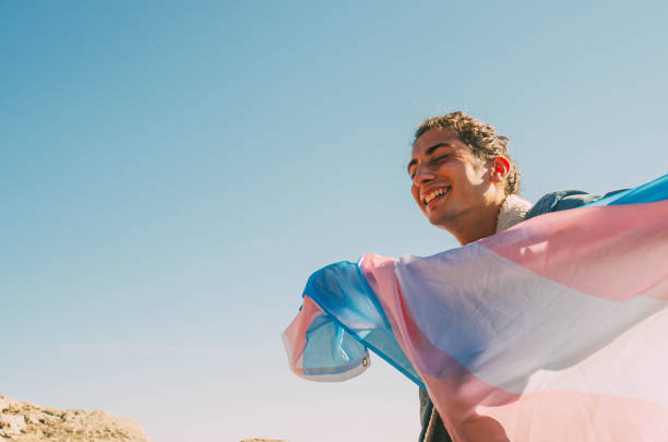 istock Happy Young Man Smiling While Holding Transgender LGTB Flag With Open Arms and Blue Sky Background 1459930351