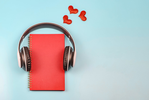 Top view or flat lay of red notebook covered with headphones on blue background decorated with red glitter hearts , copy space. Audio book, podcast, .love diary, valentine's day.