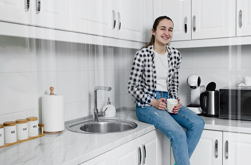 A happy Serbian female sitting on kitchen furniture and holding a coffee cup