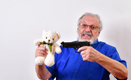 Old man with blue shirt and bow tie threatening to shoot a white toy bear with a 9mm automatic pistol