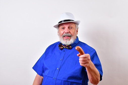 A man wearing a blue shirt, bow tie, and white hat is pointing a sausage at the camera