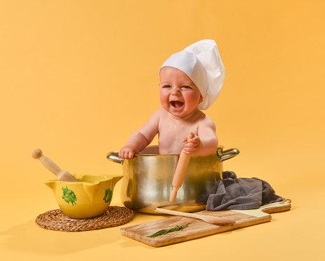 A smiling Caucasian baby tucked inside a pot with chef's hat and cooking utensils on yellow background