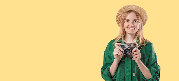 Young blonde girl holding vintage camera looking at the camera on a yellow background. Travel and vacation concept