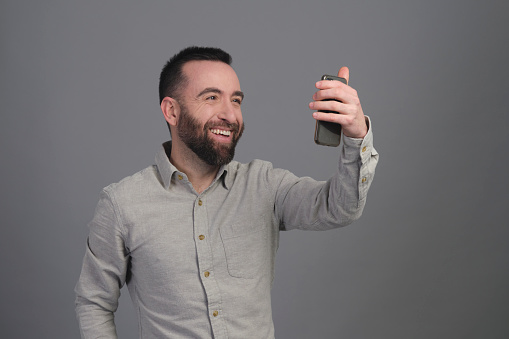 Portrait of a happy beard young man in shirt taking a selfie with his phone in a studio shot.