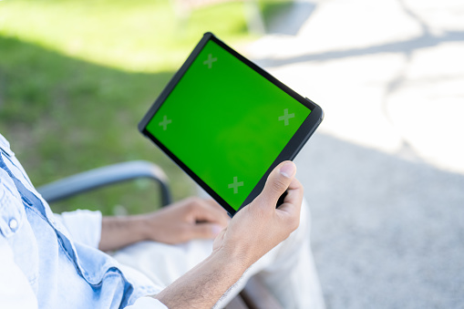 Over the shoulder view of man holding green screen of digital tablet while sitting in park.