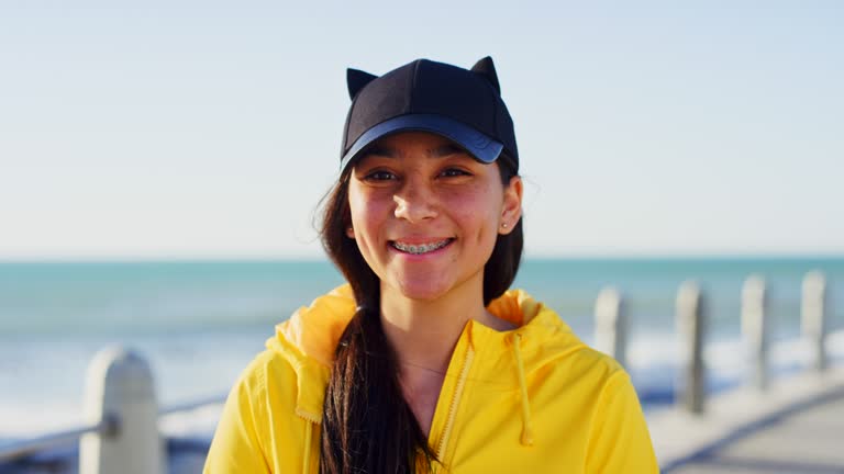 Teenager, face and smile on a beach promenade with a funny hat while on vacation in summer with happiness for travel and freedom. Portrait of a young girl outdoor in nature while happy in Miami