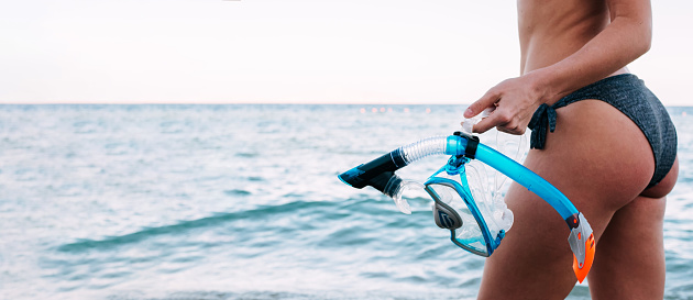 Banner of woman standing with her back to the camera holds a snorkel mask in her hand against a clear blue sky and turquoise ocean. Photo of a mask and snorkel for swimming in a pool or sea.