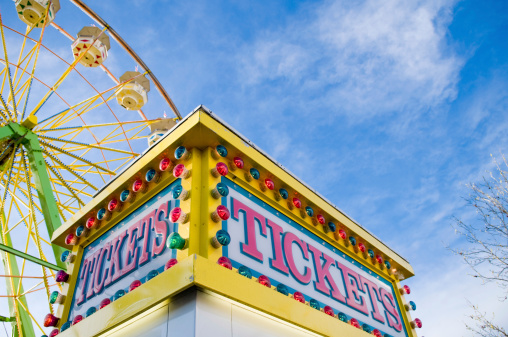 Tickets sign at county fair with Ferris wheel and light colored blue sky with white clouds in the background.