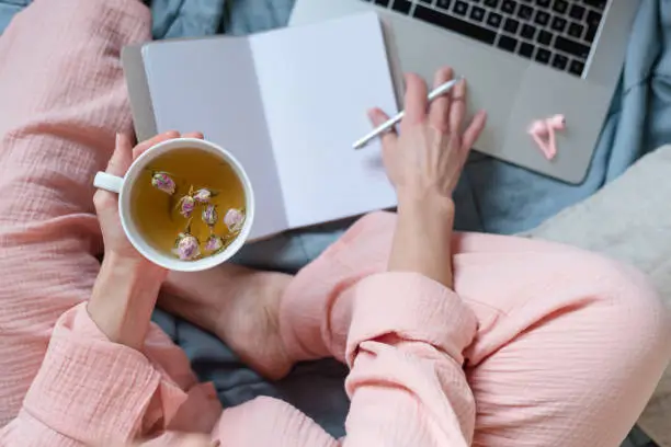 A scene in pastel colors of a woman making notes and drinking hot tea in her bed.