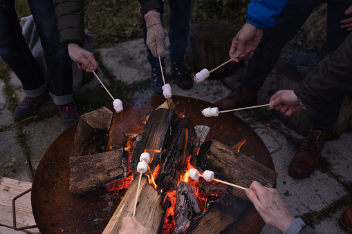 Unrecognisable personal perspective shot of a group of senior friends outdoors roasting marshmallows over a fire pit together in the North East of England. They are dressed warmly in winter.