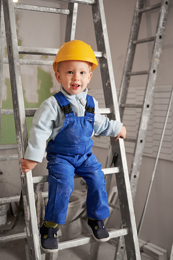 Child builder sitting on ladder in apartment under renovation. Adorable kid looking at camera and smiling, wearing safety helmet and work overalls. Home renovation and childhood concept.