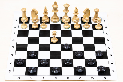 playing by different rules on the same board - black checkers and white chess figures on black white chessboard, above view of first moves of black disc and pawn (focus on the pawn)