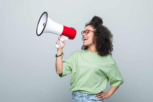 Young woman wearing green t-shirt and eyeglasses shouting into megaphone. Studio shot against grey background.