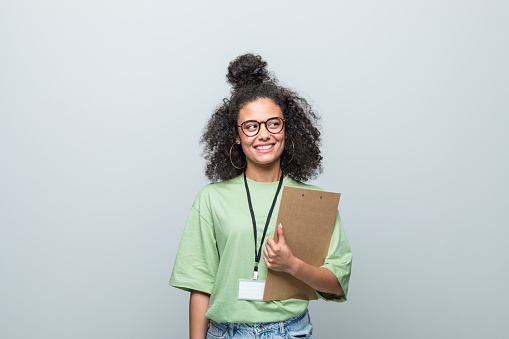 Young woman wearing green t-shirt and eyeglasses holding clipboard, looking away and smiling. Studio shot against grey background.