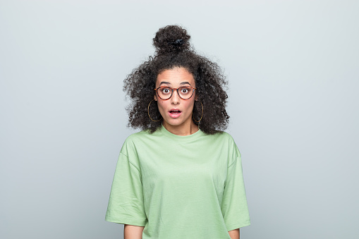 Surprised young woman wearing green t-shirt and eyeglasses looking at camera with mouth open. Studio shot against grey background.