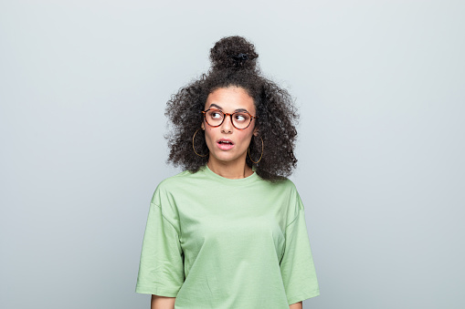 Surprised young woman wearing green t-shirt and eyeglasses looking up with mouth open. Studio shot against grey background.