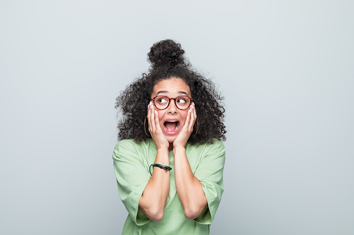 Shocked young woman wearing green t-shirt and eyeglasses holding head in hands, looking away and screaming. Studio shot against grey background.