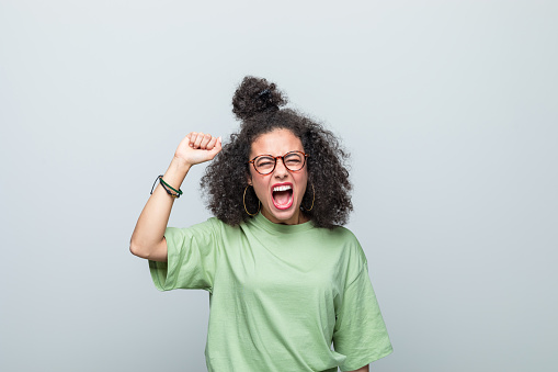 Angry young woman wearing green t-shirt and eyeglasses shouting at camera and raising fist. Studio shot against grey background.
