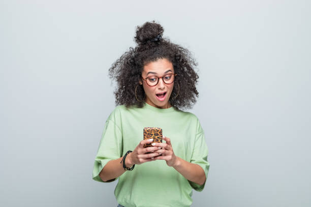 Portrait of surprised young woman using phone Excited young woman wearing green t-shirt and eyeglasses looking at smart phone with mouth open. Studio shot against grey background. black woman hair bun stock pictures, royalty-free photos & images