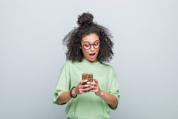 Portrait of surprised young woman using phone Excited young woman wearing green t-shirt and eyeglasses looking at smart phone with mouth open. Studio shot against grey background. phone surprise stock pictures, royalty-free photos & images