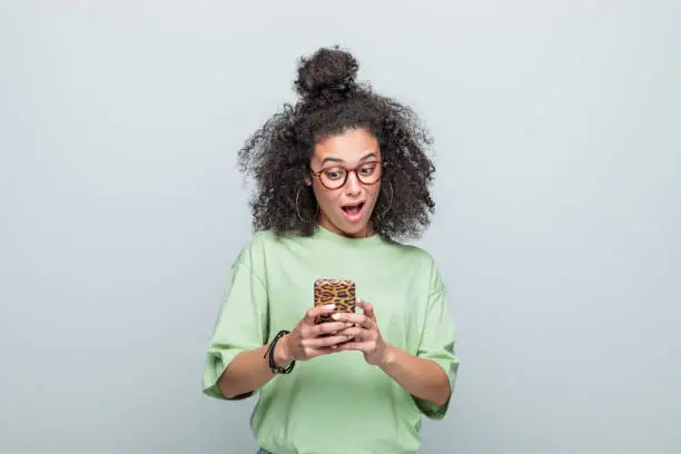 Excited young woman wearing green t-shirt and eyeglasses looking at smart phone with mouth open. Studio shot against grey background.