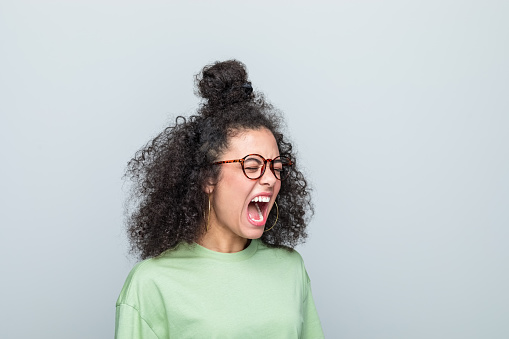 Angry young woman wearing green t-shirt and eyeglasses shouting with eyes closed. Studio shot against grey background.
