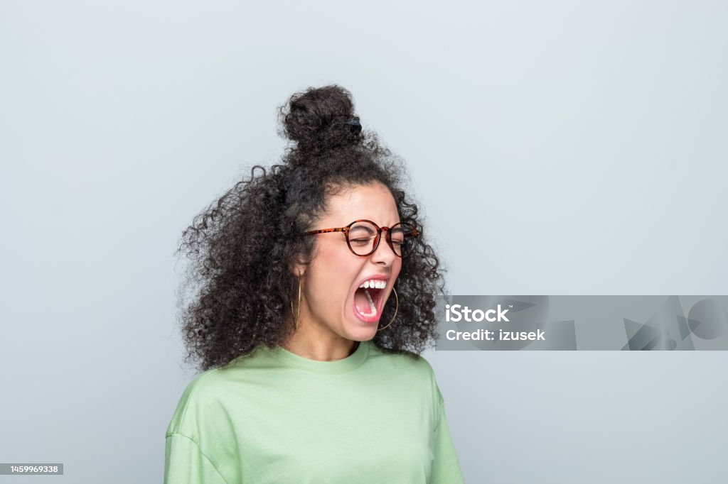 Side view of young woman shouting Angry young woman wearing green t-shirt and eyeglasses shouting with eyes closed. Studio shot against grey background. Shouting Stock Photo