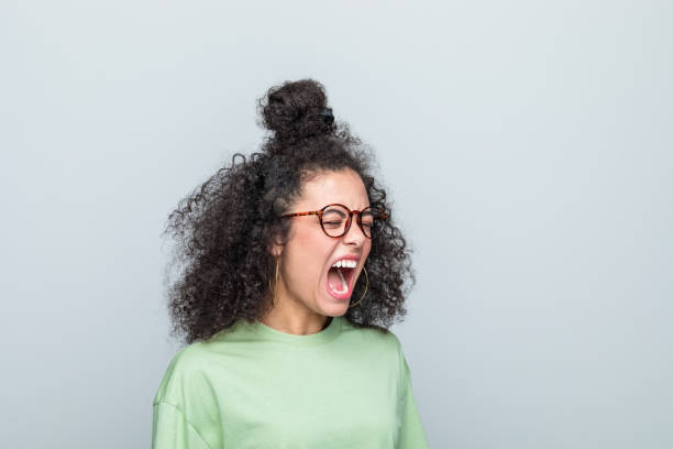 Side view of young woman shouting Angry young woman wearing green t-shirt and eyeglasses shouting with eyes closed. Studio shot against grey background. shouting stock pictures, royalty-free photos & images