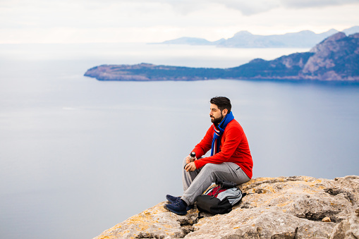 Handsome hiker sitting on a rock in front of the amazing north coast of Mallorca during the sunrise with hot tea and a thermos flask. Color editing. Part of a series.