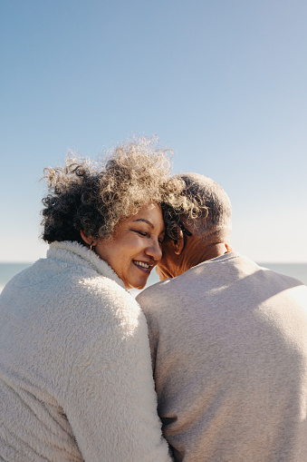 Back view of a happy senior woman smiling while sitting in front of the ocean with her husband. Retired elderly couple spending some quality time together at the beach.