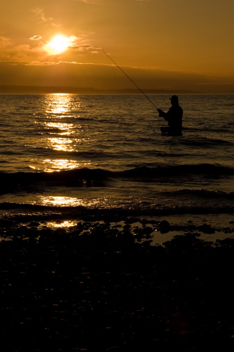 A lone silhouetted man fishing while standing on the shore of Puget Sound at Picnic Point while the sun sets over the Olympic Mountains.