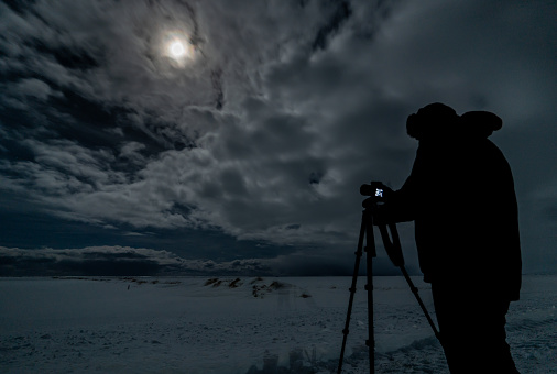 Silhouette of a photographer, photo camera and tripod on a totally snowy esplanade with the full moon illuminated the cloudy night sky