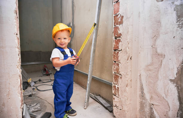 Happy boy construction worker holding measuring tape and smiling while standing in apartment under renovation. Cheerful kid with measuring tool wearing safety helmet and work overalls.
