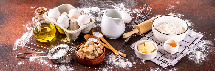 Baking ingredients background. Cooking ingredients (flour, eggs, milk, brown sugar, butter) with utensils on dark brown background top view copy space
