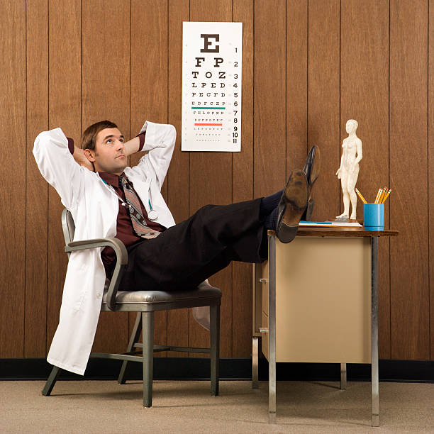Mid-adult Caucasian male doctor at desk reclining with hands behind head and feet on desk.
