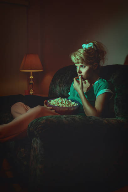 ritratto di stile di vita vintage di una giovane donna bionda alla fine degli anni '80, primi anni '90, arredamento d'interni, mangiare popcorn e rilassarsi su un divano mentre si guarda la televisione al buio - capelli cotonati foto e immagini stock