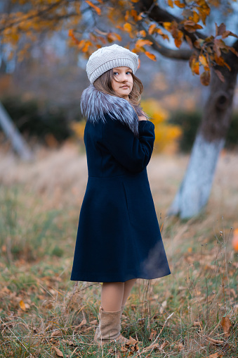 A little girl of 7 years old in a blue coat and hat is looking at the camera in the park