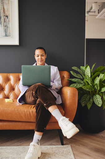Portrait of confident multiracial LGBTQ mid adult woman sitting on leather couch using laptop in retro office space
