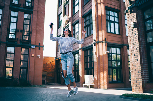 Carefree hipster girl enjoying favourite music podcast during free time at city street jumping outdoors, joyful female teenager in electronic earphones listening audio set from mobile application