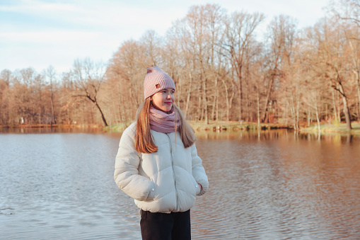 Fashionable stylish teen girl model in white jacket posing at fall park with lake outdoor, looking away. Portrait young lady standing in autumn leisure. Child youth style concept. Copy space for ad
