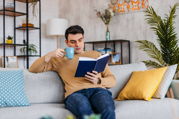 istock Young Man Reading A Book And Drinking Coffee On The Sofa 1460004832