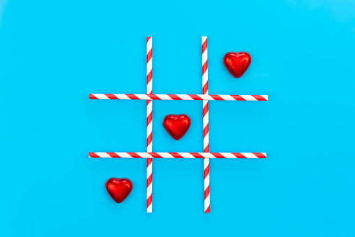 Heart shaped chocolates in red foil on blue background, tic-tac-toe game, creative concept, flat lay.