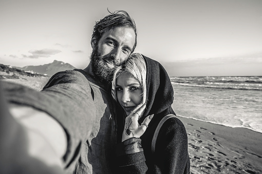 Traveling couple in love taking selfie in a deserted beach at sunset - Woman with magnetic gaze and her hipster boyfriend having fun while looking camera for selfie - Black and white editing