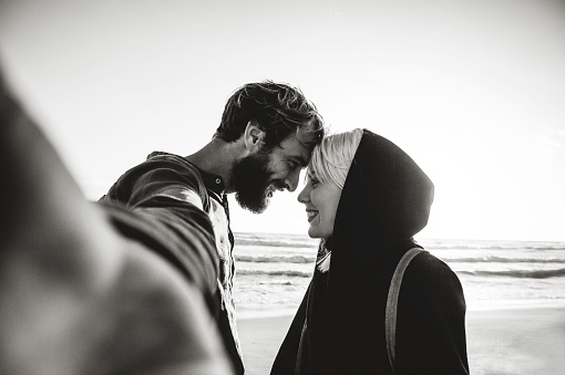 Traveling couple in love taking selfie on the deserted beach at sunset - Woman and her hipster boyfriend having fun and  looking into each other eyes while they taking a selfie - Black and white