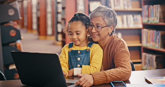 Laptop, child and grandmother reading in library for learning, education and research together. Senior woman teaching girl kid or family while streaming online, ebook subscription and learn knowledge