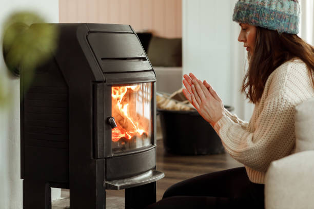 A woman sitting by a fireplace to warm up Warming up by the fireplace. Safing energy on heating. Young woman sitting at home by the fireplace and warming her hands, she is wearing white woollen sweater. Cold houses in Europe concept. Energy crisis. wood burning stove stock pictures, royalty-free photos & images