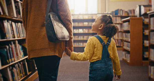istock Library, family and child education, support and holding hands for learning, teaching and bookshelf knowledge. People, mother and smart black girl in search or looking for books, story or language 1460009934