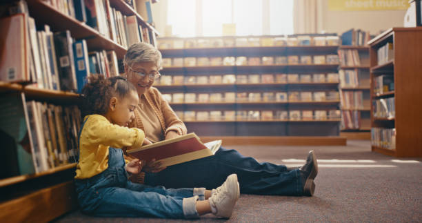 bibliothek, lernen und großmutter mit enkelkind auf dem boden mit buch zum lesen, geschichten und spaß zusammen. bildung, kinderentwicklung und oma mit mädchen, geschichtenerzählen und unterrichten während der bindung - grandparent family reading inside of stock-fotos und bilder