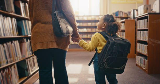 istock Library, help child and woman search books, learning and education support and holding hands for fun hobby. Kindergarten girl and family or people by bookshelf for back to school and mind development 1460011110