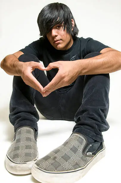 Teen boy dressed in black sitting on floor making heart shape with hands expressing love